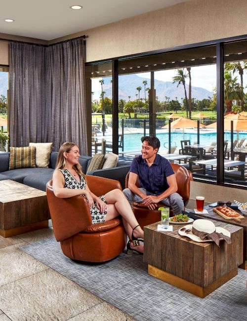 A couple sits in a cozy lounge area, enjoying drinks and snacks with a view of a swimming pool and mountains outside the window.