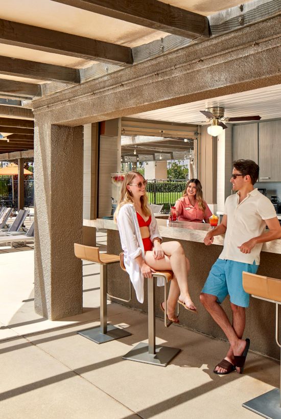 People are at an outdoor bar with drinks, under a shaded area. Orange umbrellas and loungers are visible near a pool in the background.