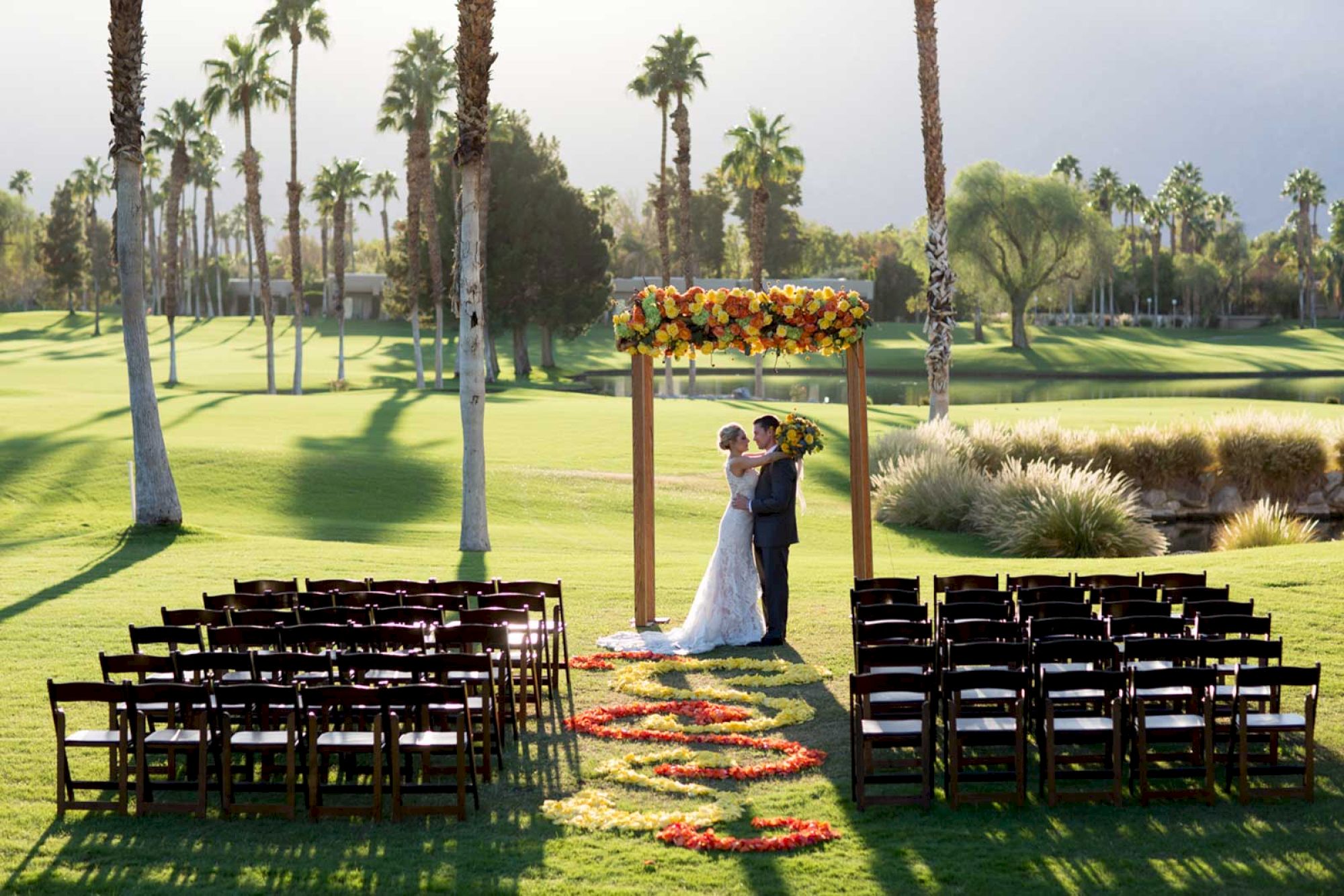 A couple stands under a floral arch on a lawn with palm trees, surrounded by arranged chairs for a wedding ceremony.