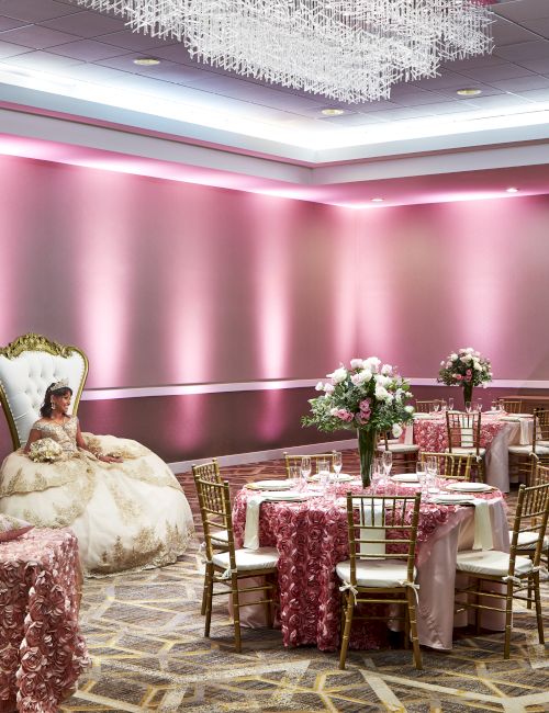 A lavishly decorated pink event hall with elegant tables and a woman in a formal dress seated on a large chair.