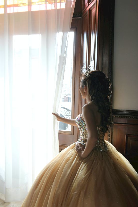 A person in a glamorous gown stands by a window, curtains drawn slightly back, gazing outside, with elegant wood paneling in the background.