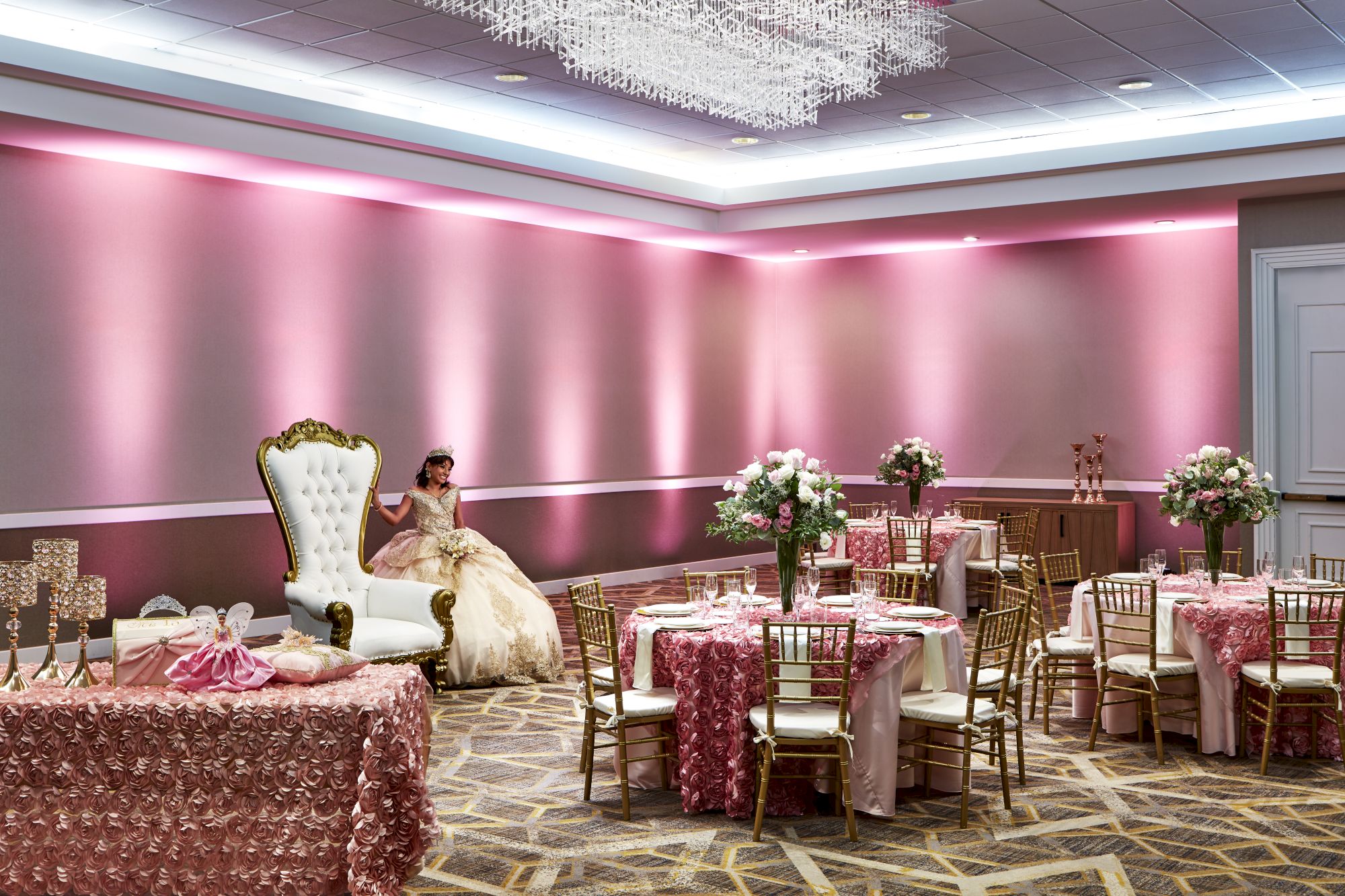 A ballroom with pink decor, a throne-like chair, floral centerpieces, and a person in formal attire seated on the chair.