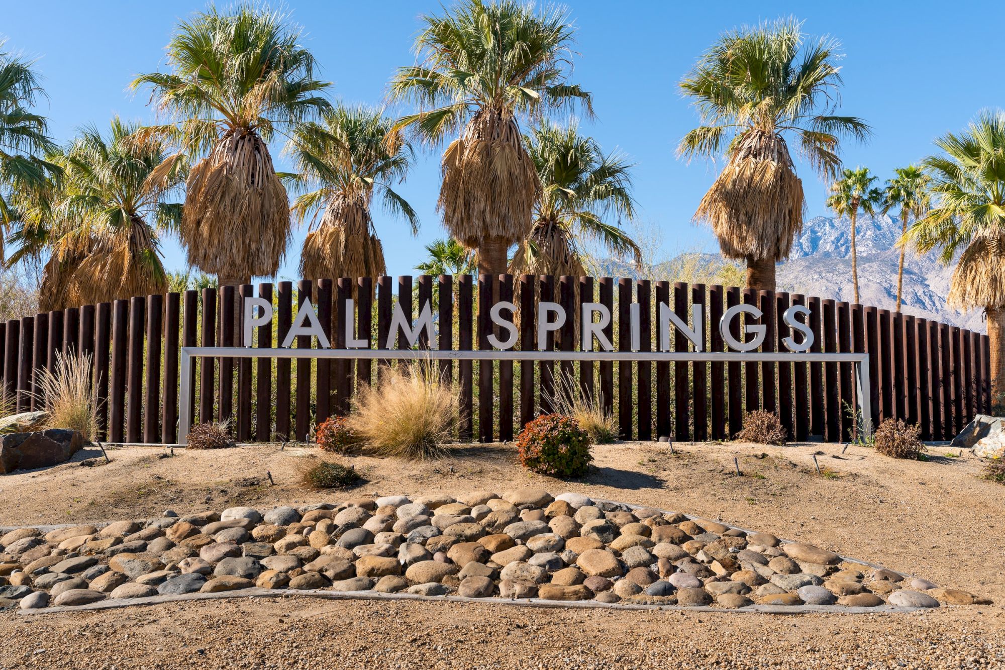 The image shows a "Palm Springs" sign with palm trees and stones in front, under a clear blue sky.