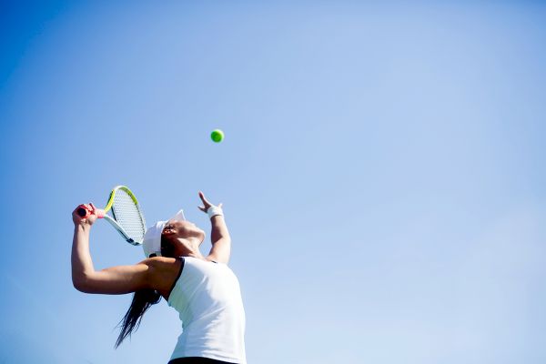 A person is serving a tennis ball under a clear blue sky, wearing a white tank top and headband, holding a tennis racket.