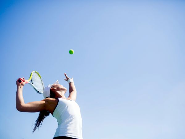 A person is serving a tennis ball under a clear blue sky, wearing a white tank top and headband, holding a tennis racket.