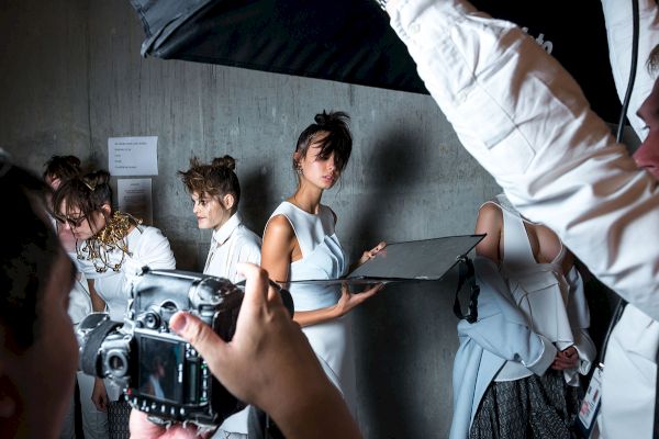 Models in white outfits are being photographed backstage, with lighting and cameras capturing the scene.