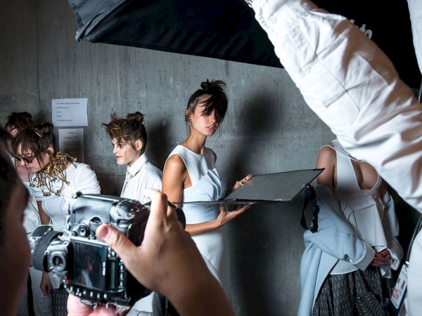 Models in white outfits are being photographed backstage, with lighting and cameras capturing the scene.
