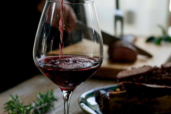 A glass of red wine is being poured on a table with a person slicing food in the background. Fresh herbs are also visible on the table.