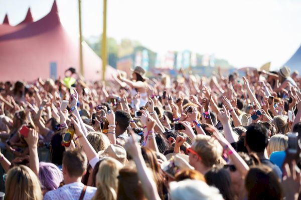 A large crowd at an outdoor music festival, with many people raising their hands and enjoying the event under a sunny sky.