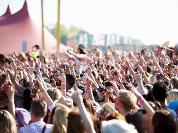 A large crowd at an outdoor music festival, with many people raising their hands and enjoying the event under a sunny sky.