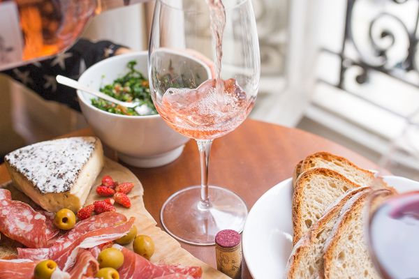 A charcuterie board with meats, olives, and cheese, bread, and two glasses of rosé wine are served near a window.