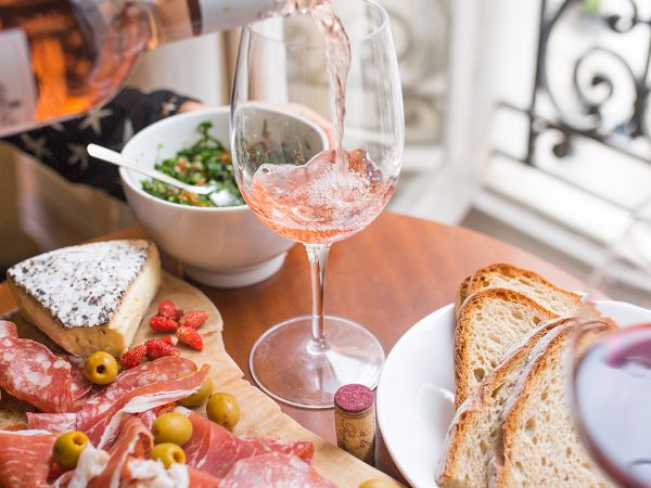 A charcuterie board with meats, olives, and cheese, bread, and two glasses of rosé wine are served near a window.