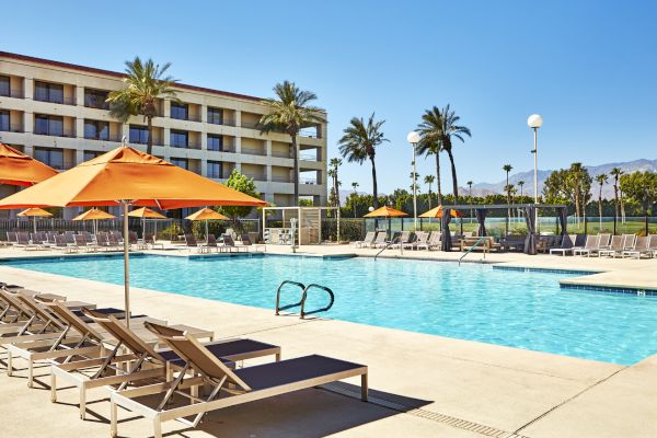A sunny poolside scene with orange umbrellas, lounge chairs, and a multi-story building surrounded by palm trees.