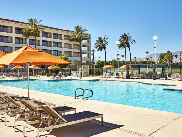A sunny poolside scene with orange umbrellas, lounge chairs, and a multi-story building surrounded by palm trees.