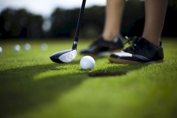 A golf club near a ball on the grass, with a person's legs and multiple golf balls in the background.