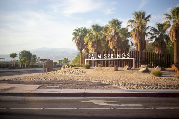 A sign for "Palm Springs" surrounded by palm trees and desert landscape, with mountains in the background.