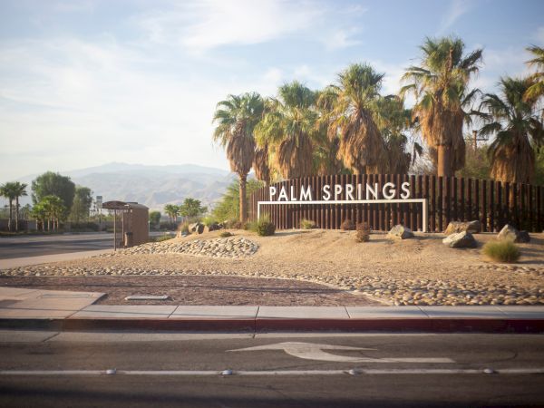 A sign for "Palm Springs" surrounded by palm trees and desert landscape, with mountains in the background.