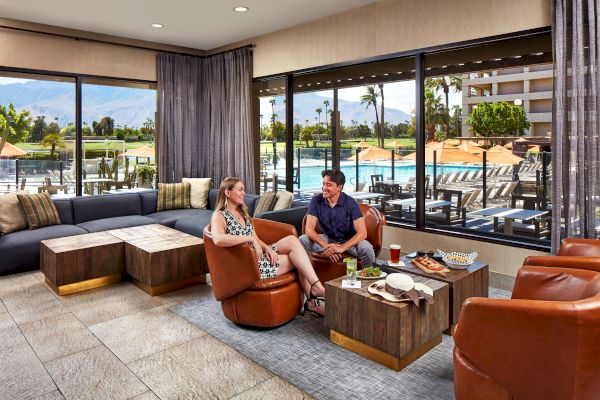 Two people sitting and talking in a lounge with a view of a pool and mountains through large windows.