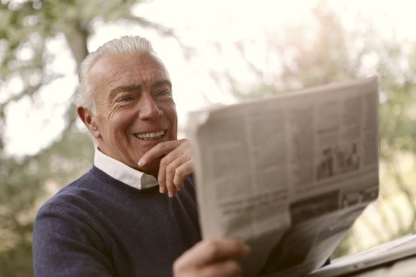 An older man is smiling while reading a newspaper outdoors, with trees blurred in the background.