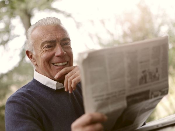 An older man is smiling while reading a newspaper outdoors, with trees blurred in the background.