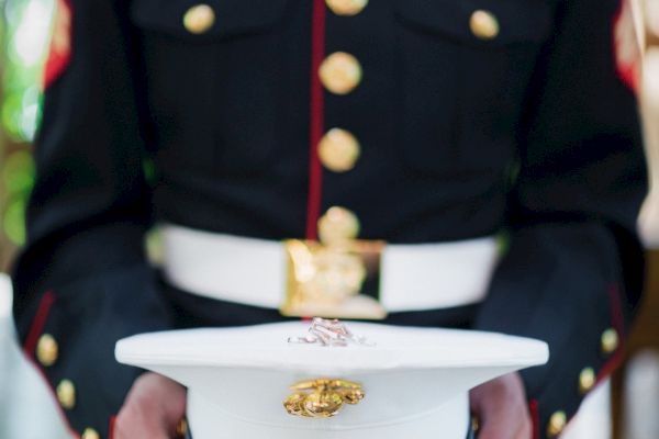 A person in a decorated military uniform holds a white hat with gold emblems, set against a blurred outdoor background.