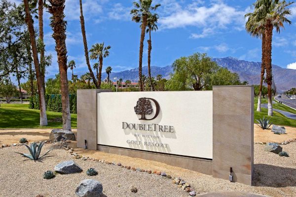 A sign for DoubleTree by Hilton Golf Resort in a desert landscape with palm trees and mountains in the background.