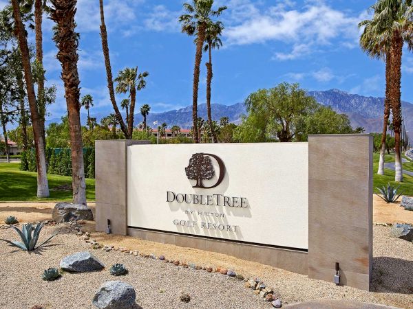 A sign for DoubleTree by Hilton Golf Resort in a desert landscape with palm trees and mountains in the background.