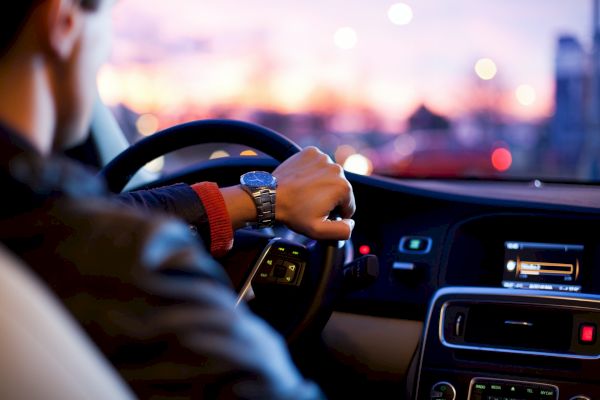A person is driving a car, with a focus on the steering wheel and dashboard at sunset.