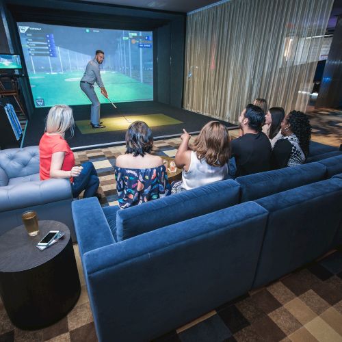 People on a sofa watching someone play golf on a simulator screen in an indoor entertainment space.
