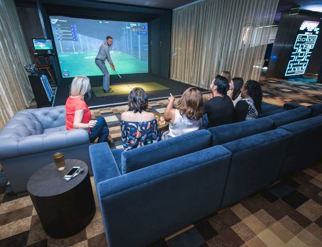 People on a sofa watching someone play golf on a simulator screen in an indoor entertainment space.
