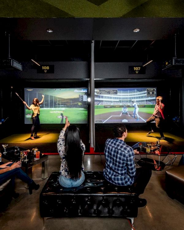 People are in an indoor entertainment venue with sports simulators, golf screens, and seating areas, engaging in activities and socializing.