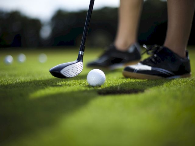 A person is golfing on a green, with a club near a golf ball, and wearing sneakers.