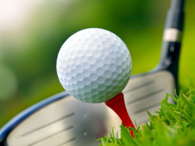 A golf ball is perched on a red tee with a golf club poised behind it, ready for a shot on a grassy course.