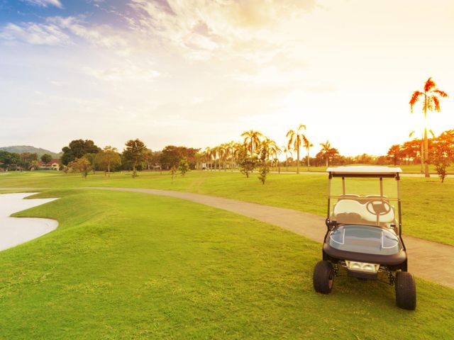 A golf course featuring a golf cart on lush green grass, surrounded by trees under a partly cloudy sky during sunset.