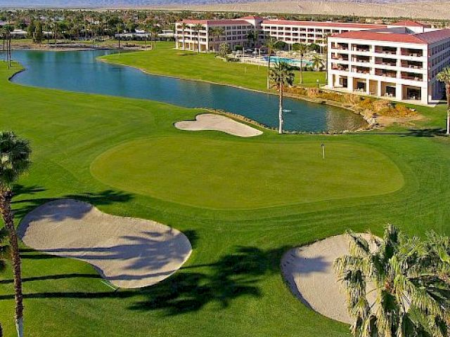 The image shows a resort with a golf course, featuring sand bunkers, a pond, and palm trees, set against distant mountains and desert.