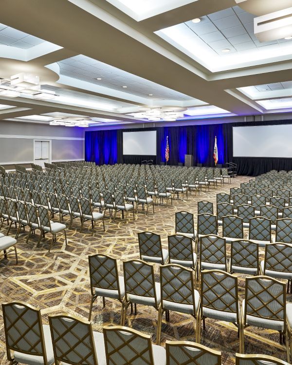An empty conference room with rows of chairs, two large screens, and blue curtains, ready for an event or presentation.