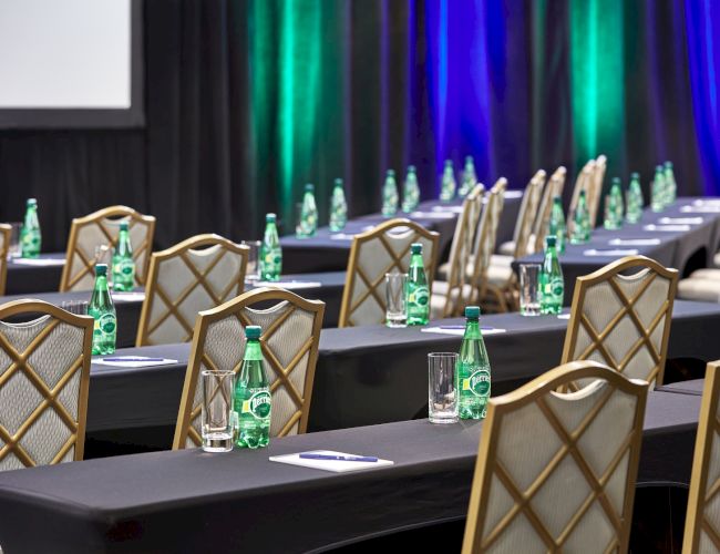 An empty conference room with rows of tables and chairs, bottles of water, notepads, and glasses, set up for a meeting or event.
