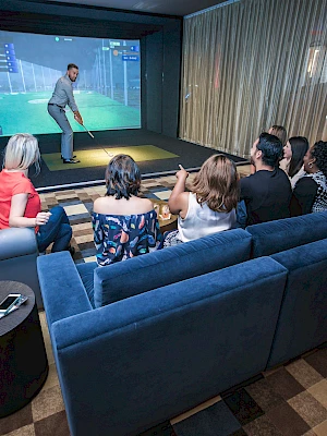 A group of people sits on couches, watching someone playing a golf simulator in a modern lounge setting.