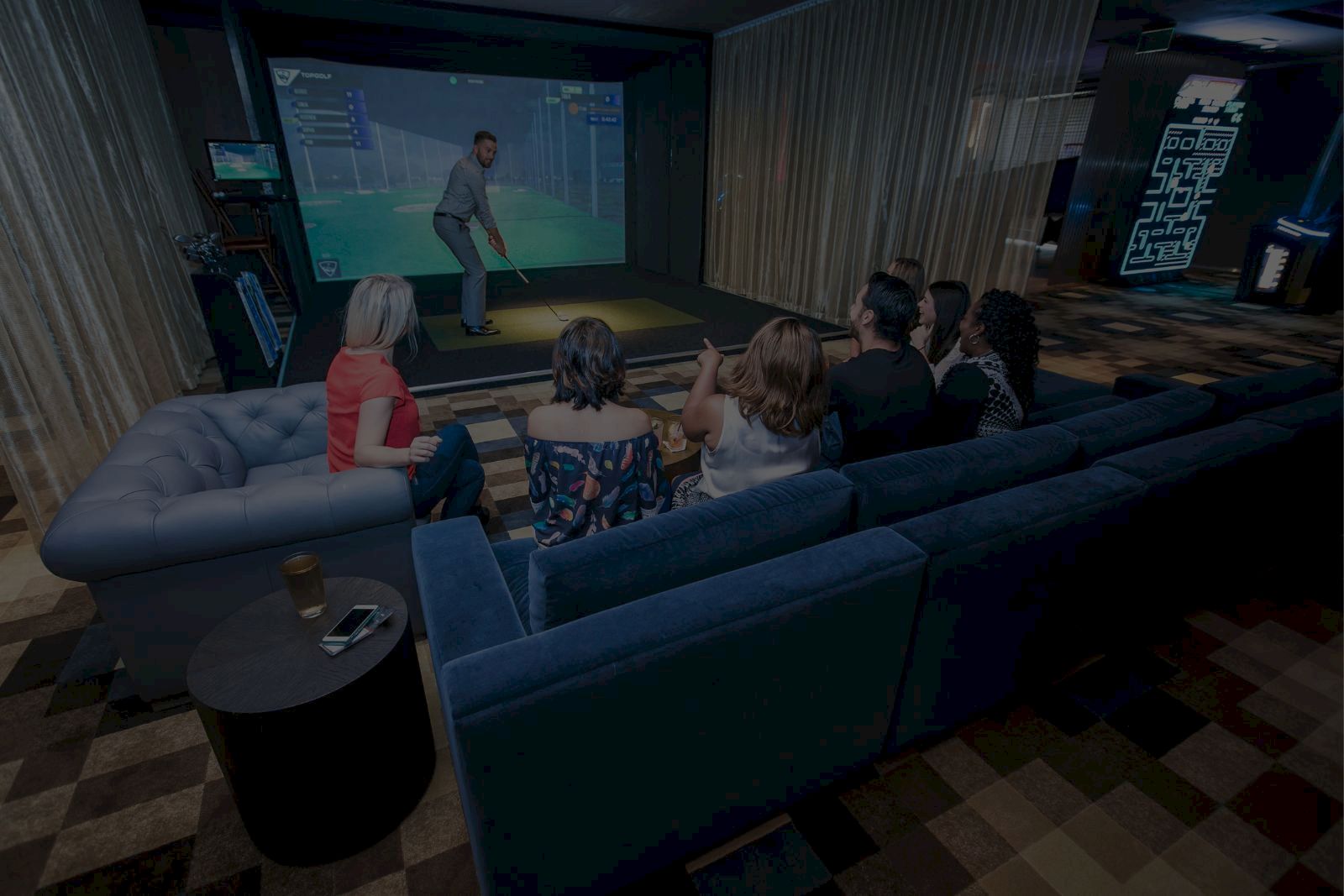 A group of people sits on couches watching someone play a golf simulator in a cozy room.