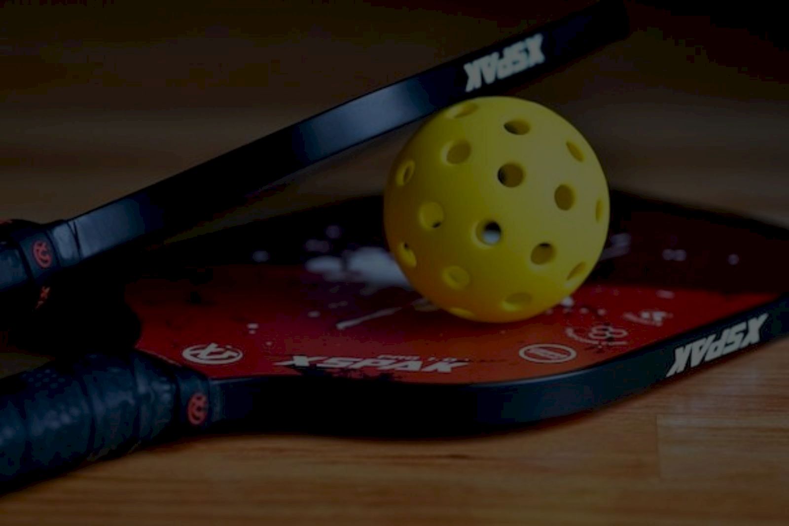 The image shows two pickleball paddles with a yellow perforated ball on a wooden surface.