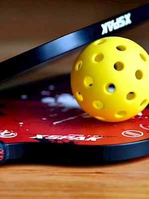 The image shows two pickleball paddles with a yellow perforated ball resting on them, placed on a wooden surface.