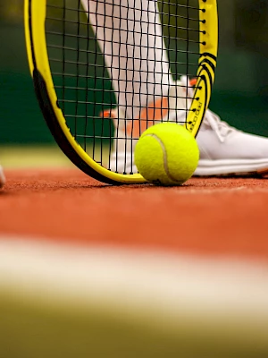 A tennis racket and ball are on a clay court next to a player's feet in white sneakers, ready for action.