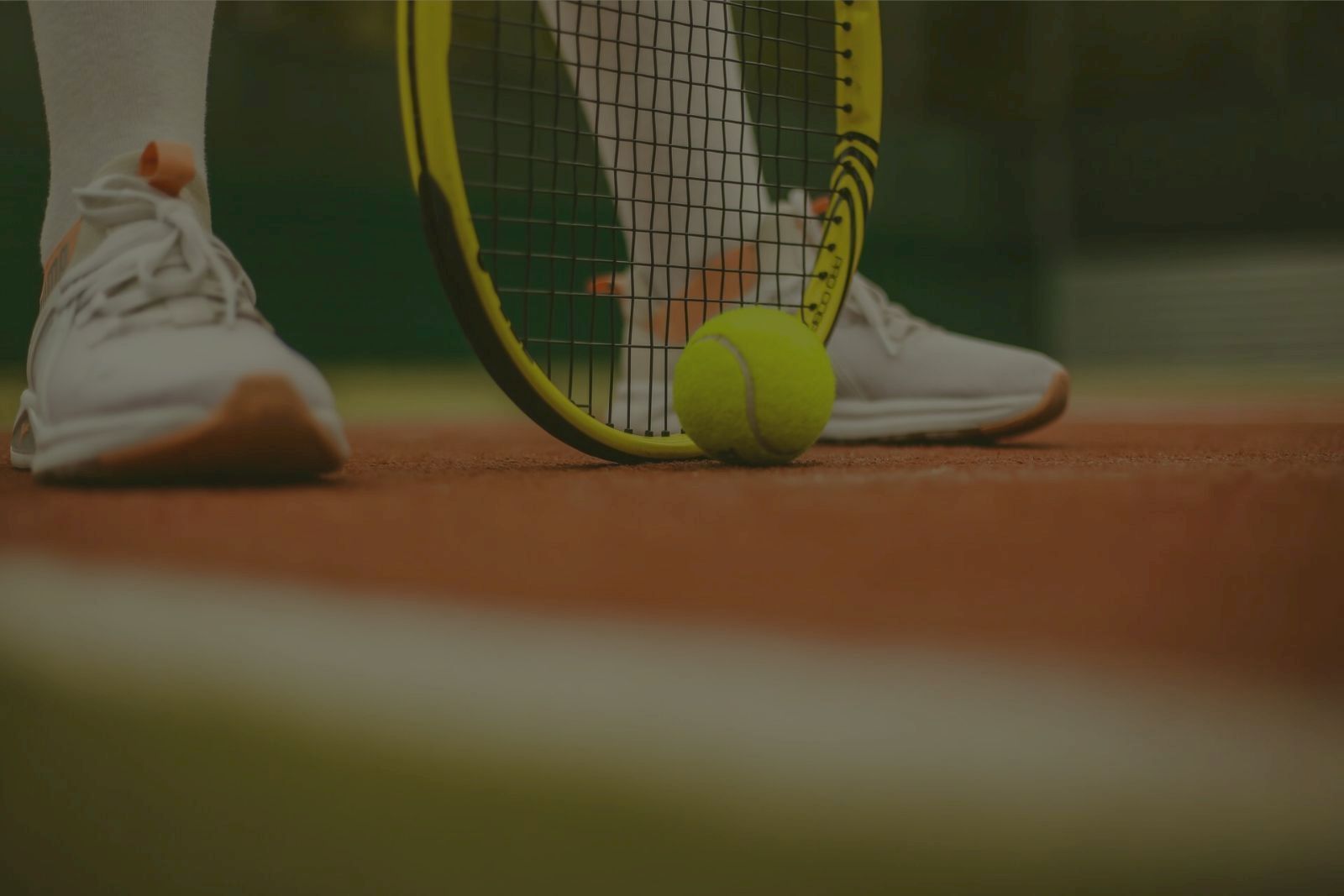 Tennis court scene with a close-up of sneakers, a racquet, and a tennis ball resting on the red surface.