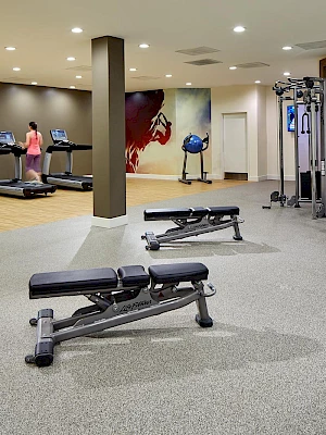 A modern gym with treadmills, weight benches, dumbbells, and fitness equipment. A person exercises on a treadmill in the background.