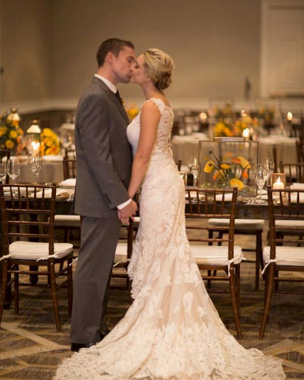 A couple in wedding attire stands together in a warmly decorated indoor setting, surrounded by tables with floral arrangements.