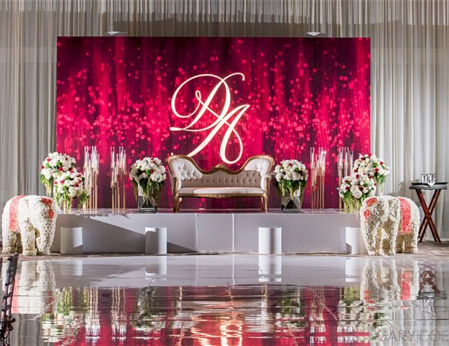 An elegant event stage with a sofa, floral arrangements, and decorative elephants, set against a red backdrop with "DA" initials.