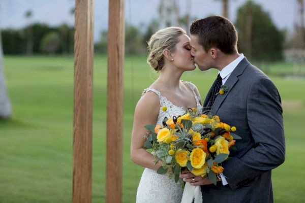 A couple in wedding attire shares a kiss outdoors, the bride holding a bouquet of yellow flowers, surrounded by a green lawn and wooden posts.