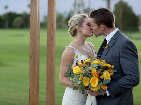 A couple in wedding attire shares a kiss outdoors, the bride holding a bouquet of yellow flowers, surrounded by a green lawn and wooden posts.