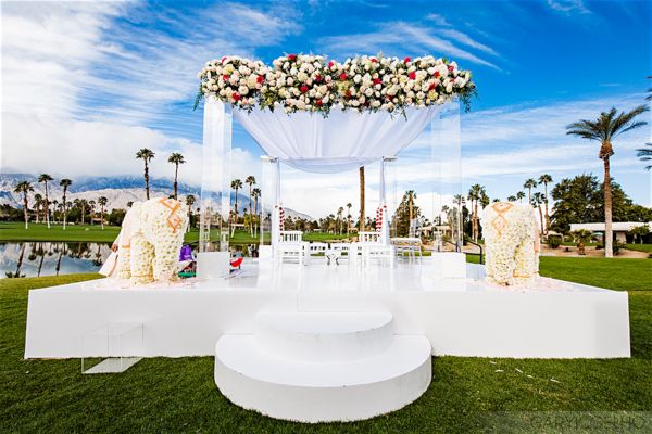 An outdoor wedding altar with floral arrangements, white drapery, and decorated elephants in a scenic setting with palm trees and a clear sky.