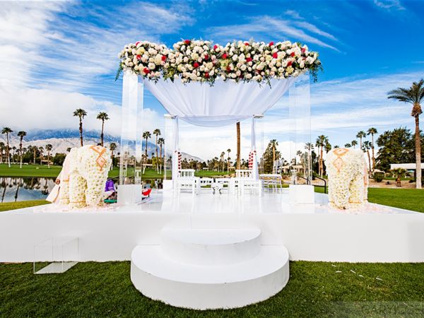 An outdoor wedding altar with floral arrangements, white drapery, and decorated elephants in a scenic setting with palm trees and a clear sky.